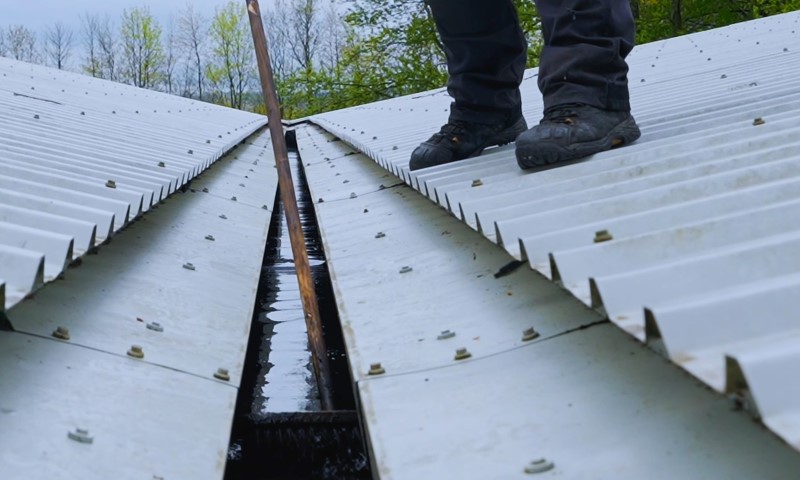 Why Roof and Gutter Maintenance Matters for Commercial Buildings 2 Person standing on a corrugated metal roof using a broom to clean a gutter