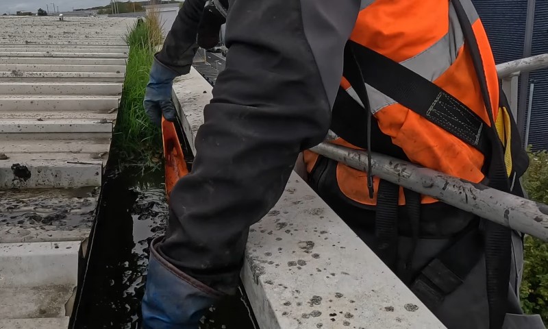 Why Roof and Gutter Maintenance Matters for Commercial Buildings 4 A person wearing gloves and an orange safety vest is scooping debris from a narrow water channel