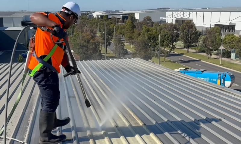 Why Roof and Gutter Maintenance Matters for Commercial Buildings 5 A worker in a hard hat and safety gear power washes a corrugated metal roof